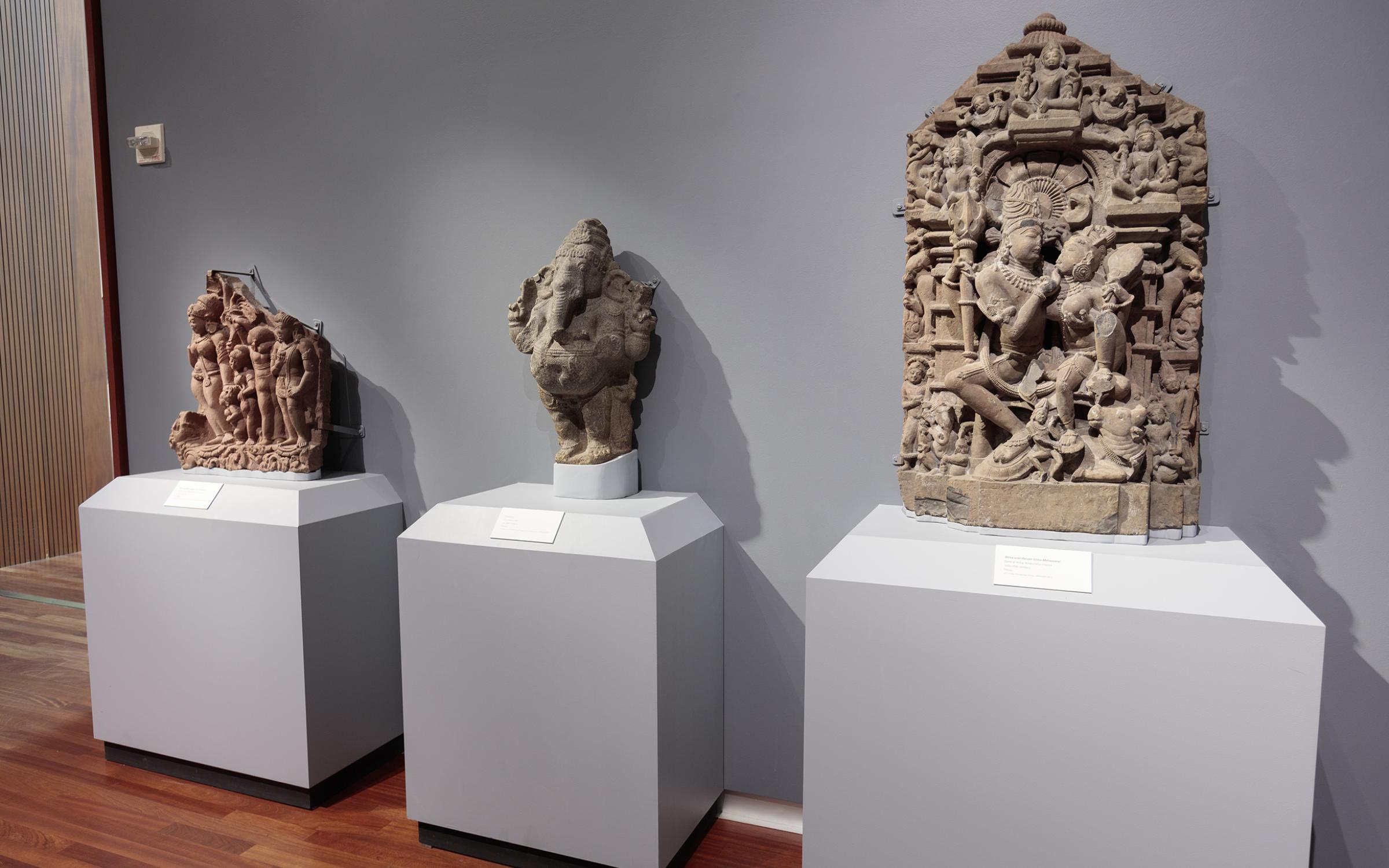 There are three intricately carved deities lined up on individual stands against a light grey wall.