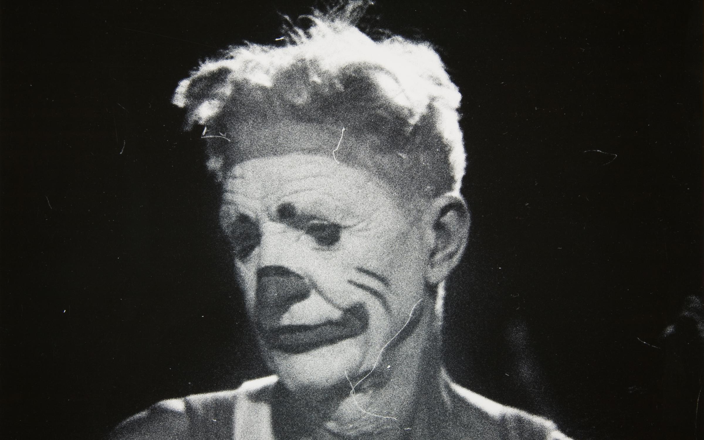 A black and white photo of figure in clown make up. The figure looks down and to the side as though sad.