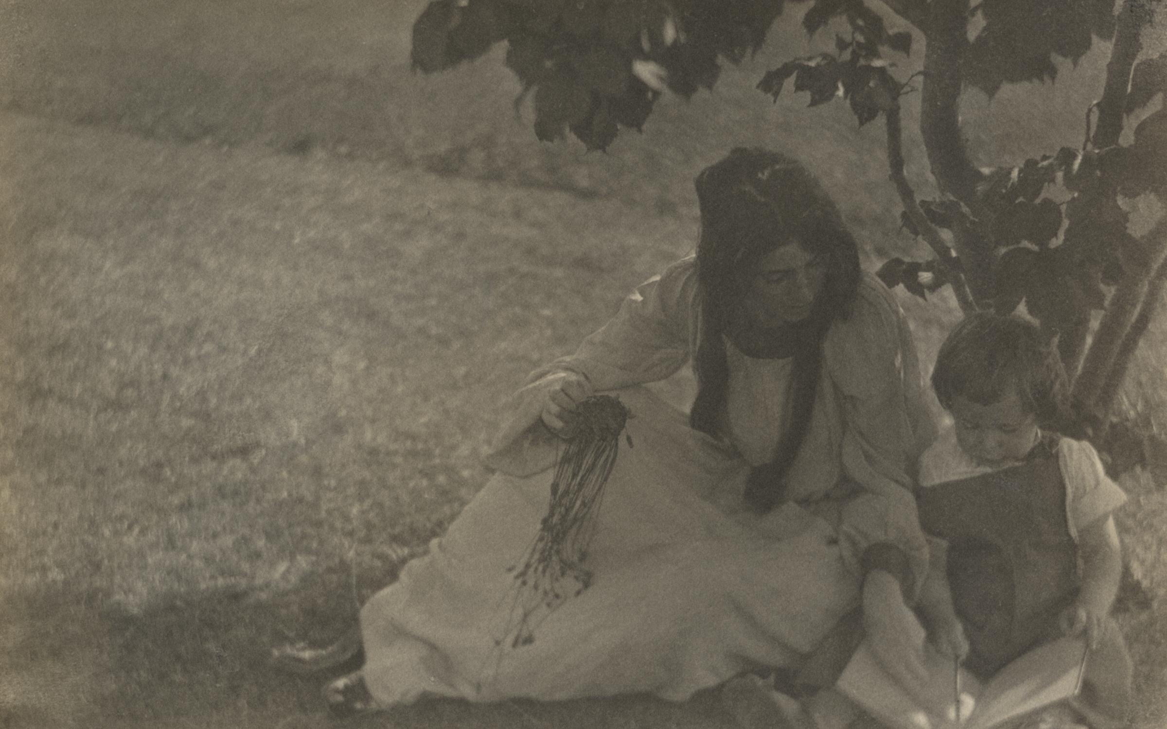 A black and white photo of a woman in a white dress sitting on the ground beneath a tree with a small child looking at a book.