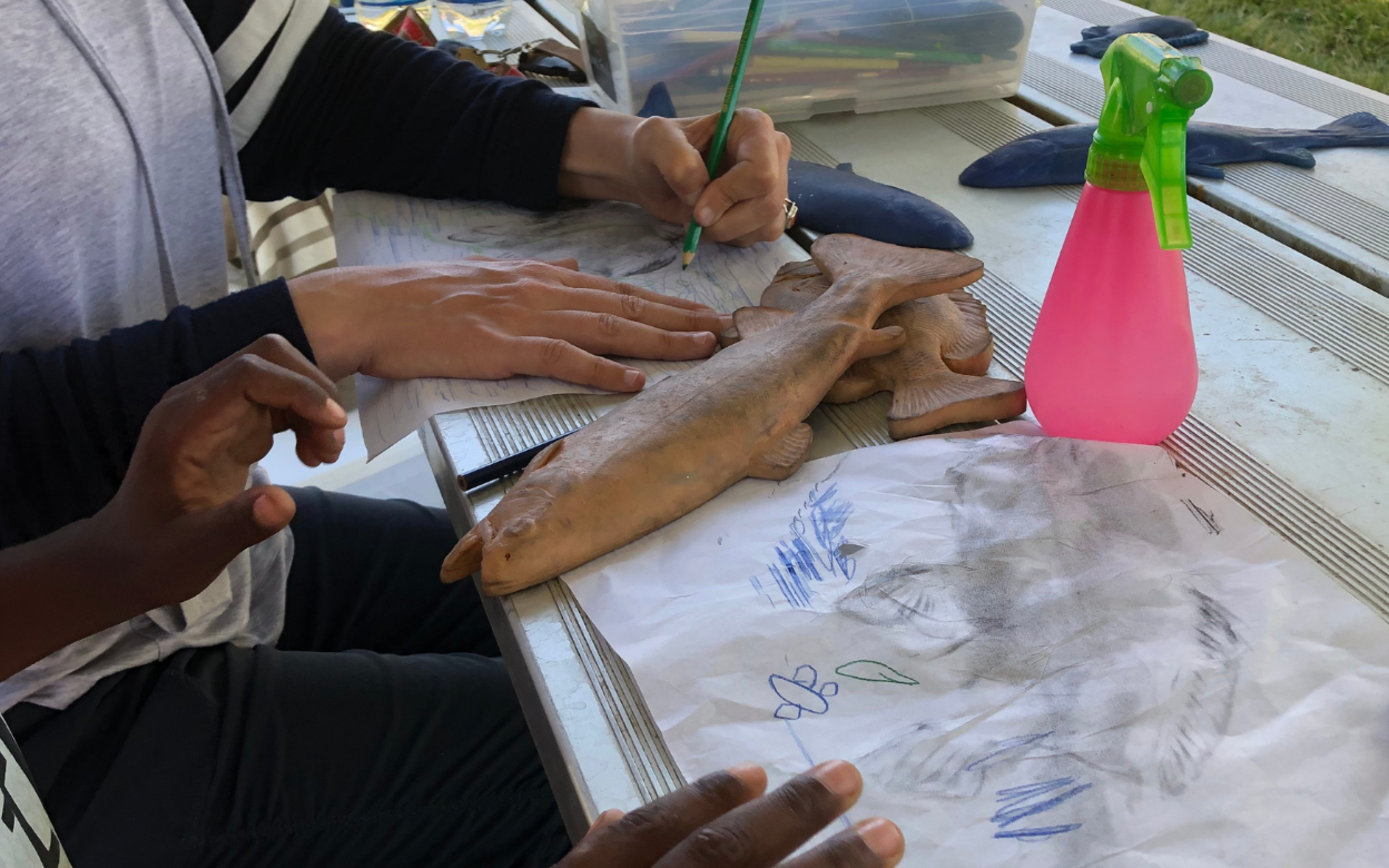 A table is busy with crafting hands. A clay fish lays next to a pencil-rubbing on paper.