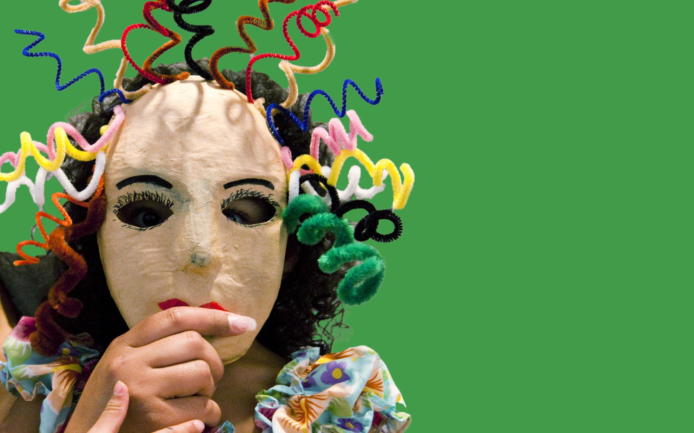 A child holds a mask with curly pipe cleaner hair to their face.