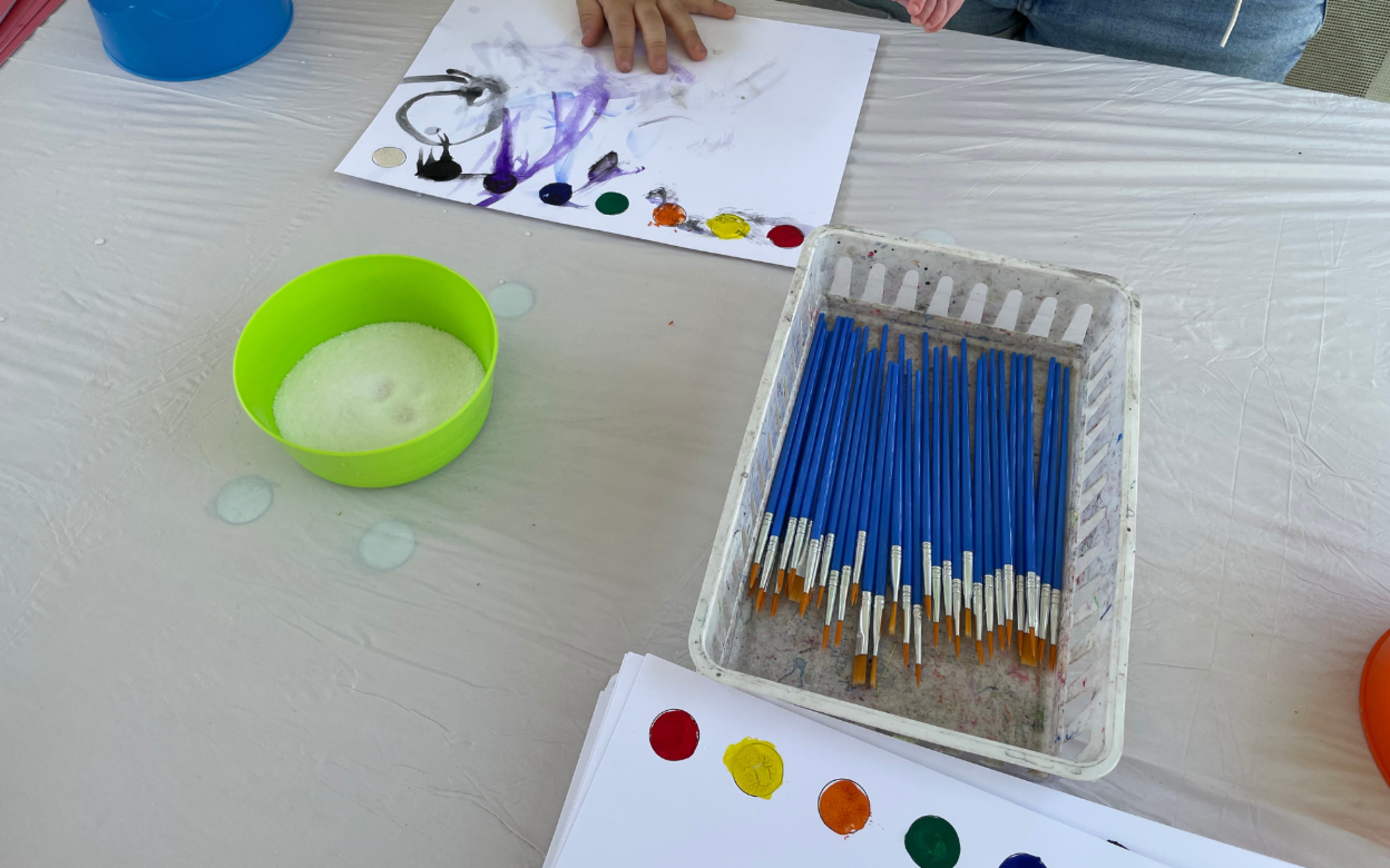 Blue Moon Festival A table with paint brushes and white paper with watercolor dots on them. At the top of the photograph a child's hand smudges the paint.