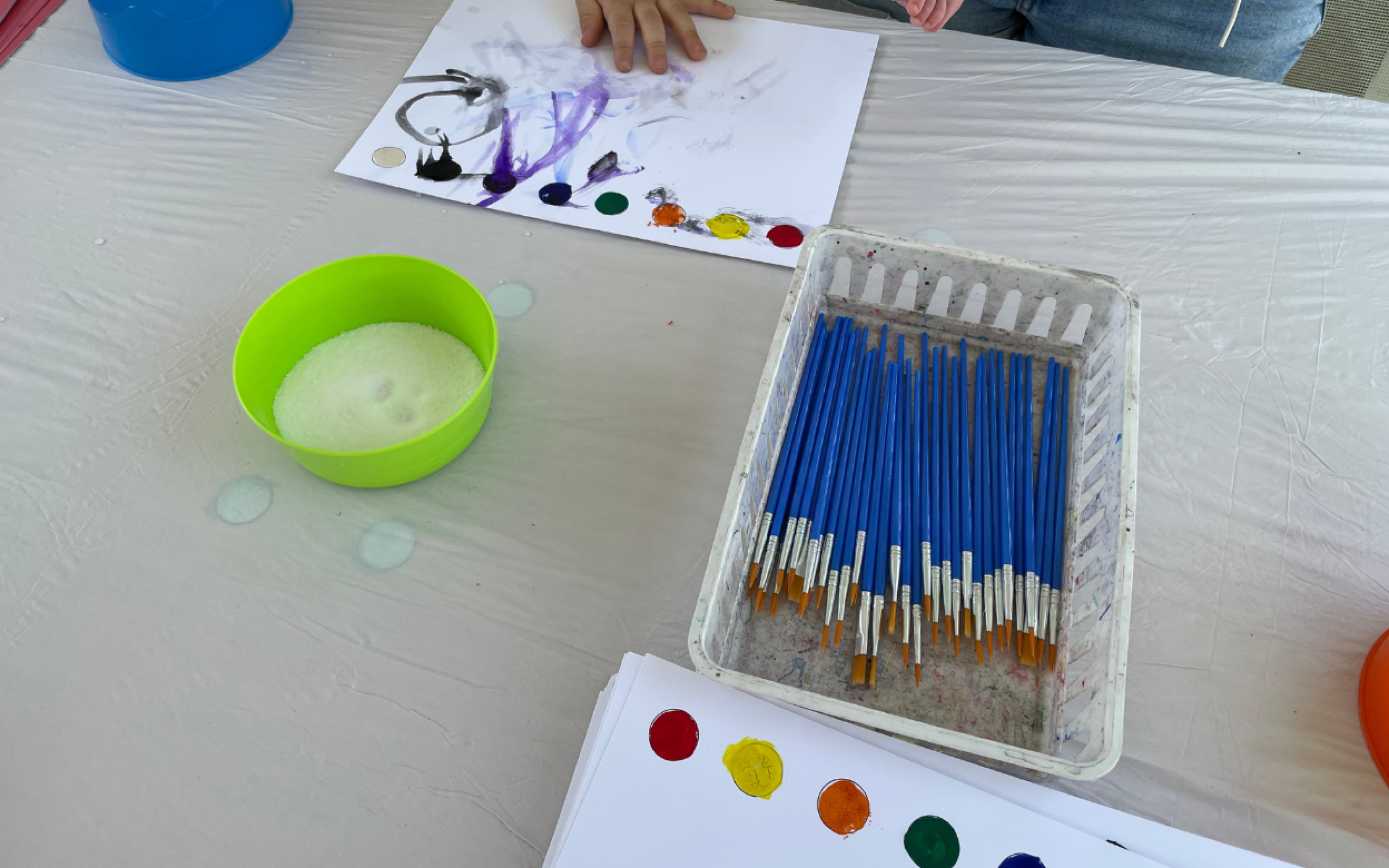 A table with paint brushes and white paper with watercolor dots on them. At the top of the photograph a child's hand smudges the paint. 