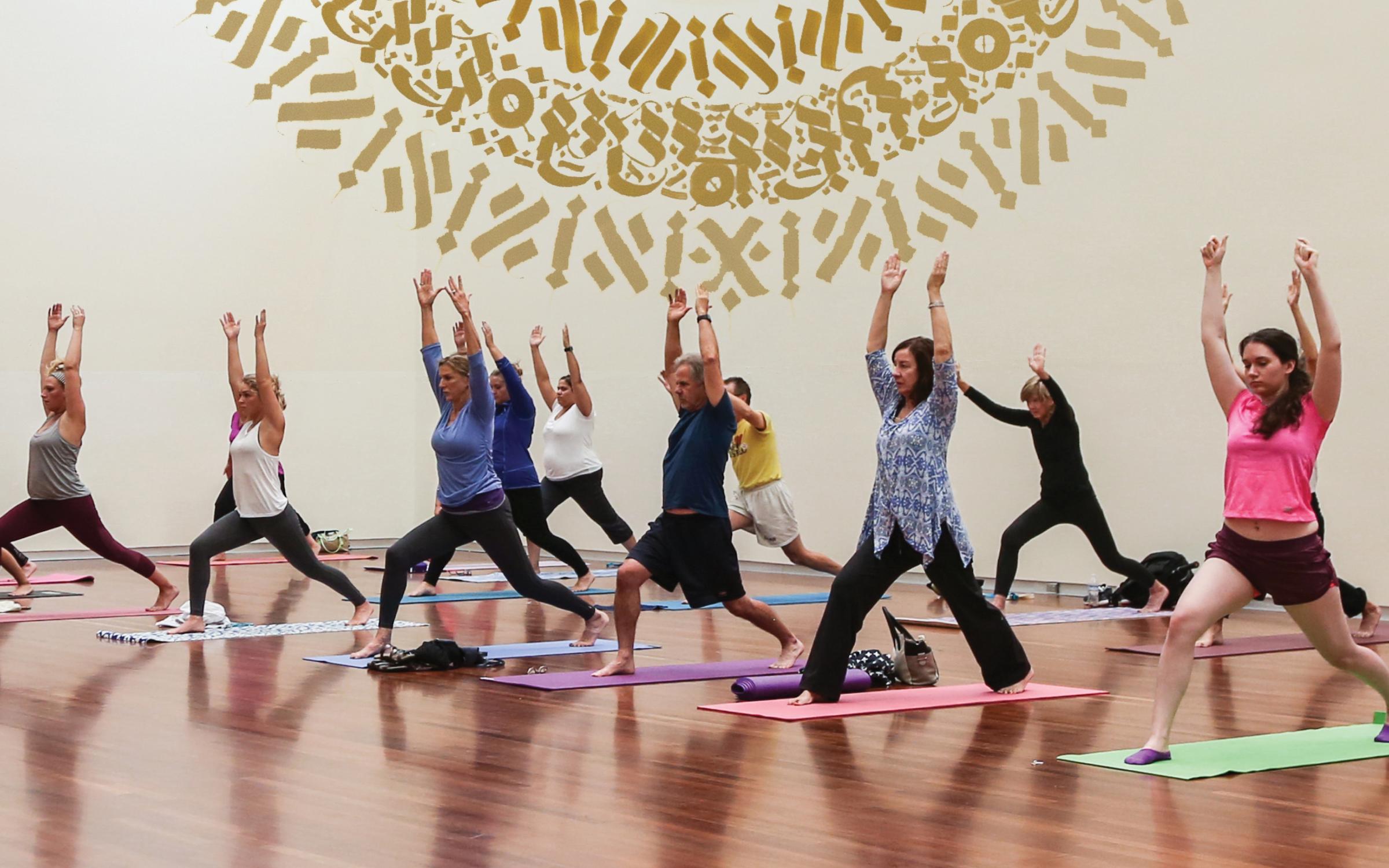 A group of people practice yoga in the UMFA Great Hall. A gold mural is in the background.