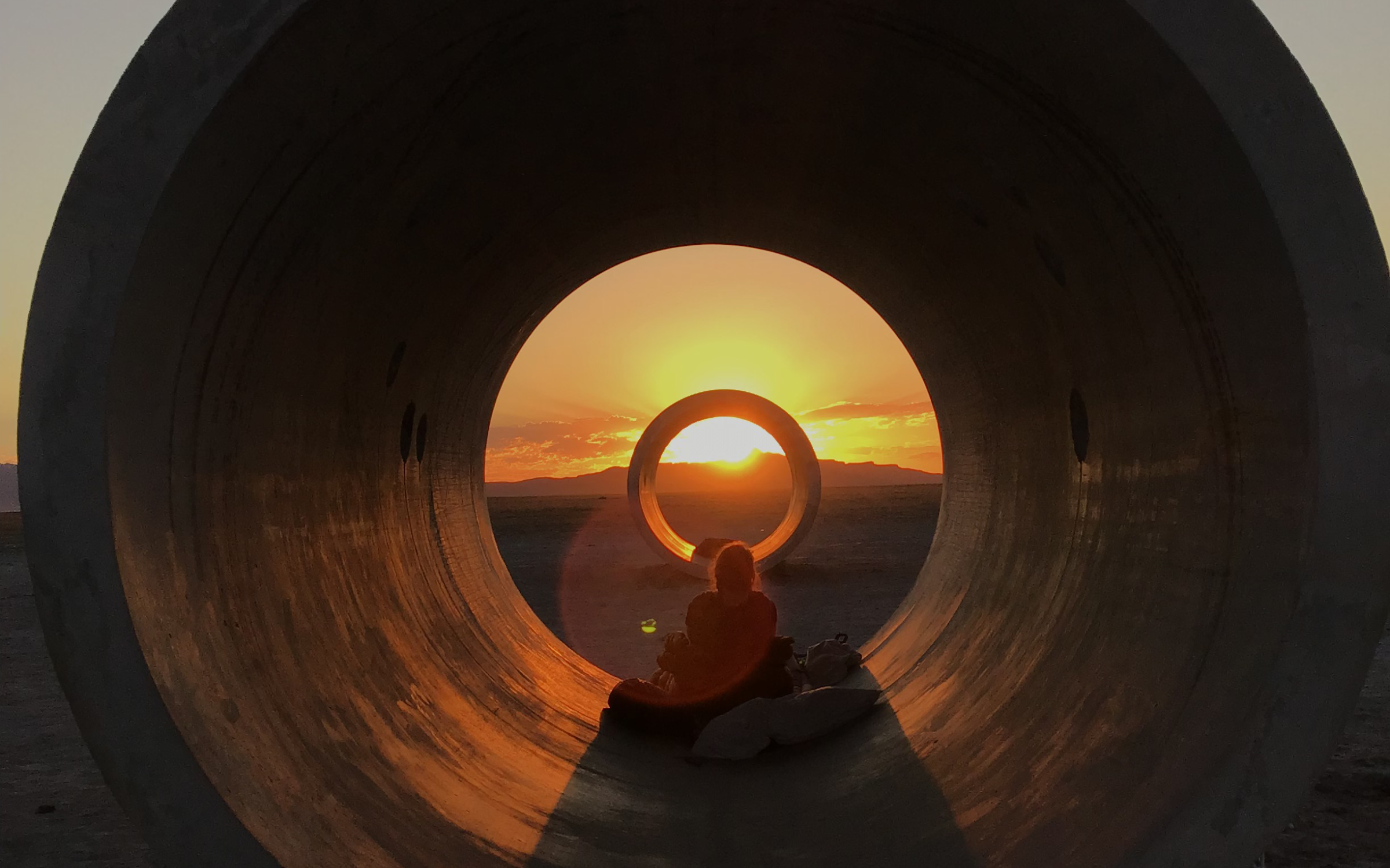 Two concrete tunnels line up with the setting sun. A person sits silhouetted between the two tunnels surrounded by warm orange light.