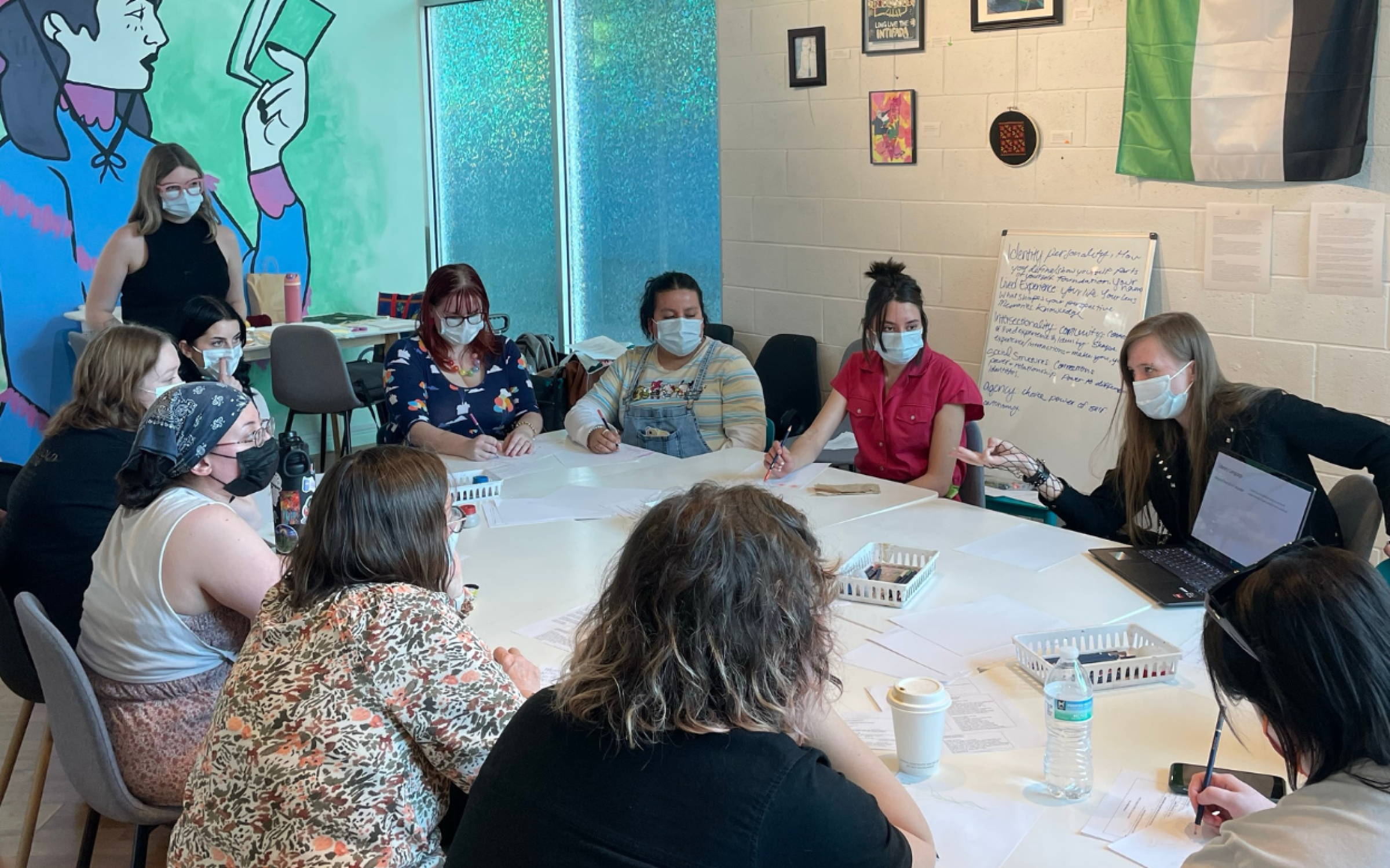 ACME Workshop A group of people in masks sit around a table. A mural of a woman reading a book is in the background.