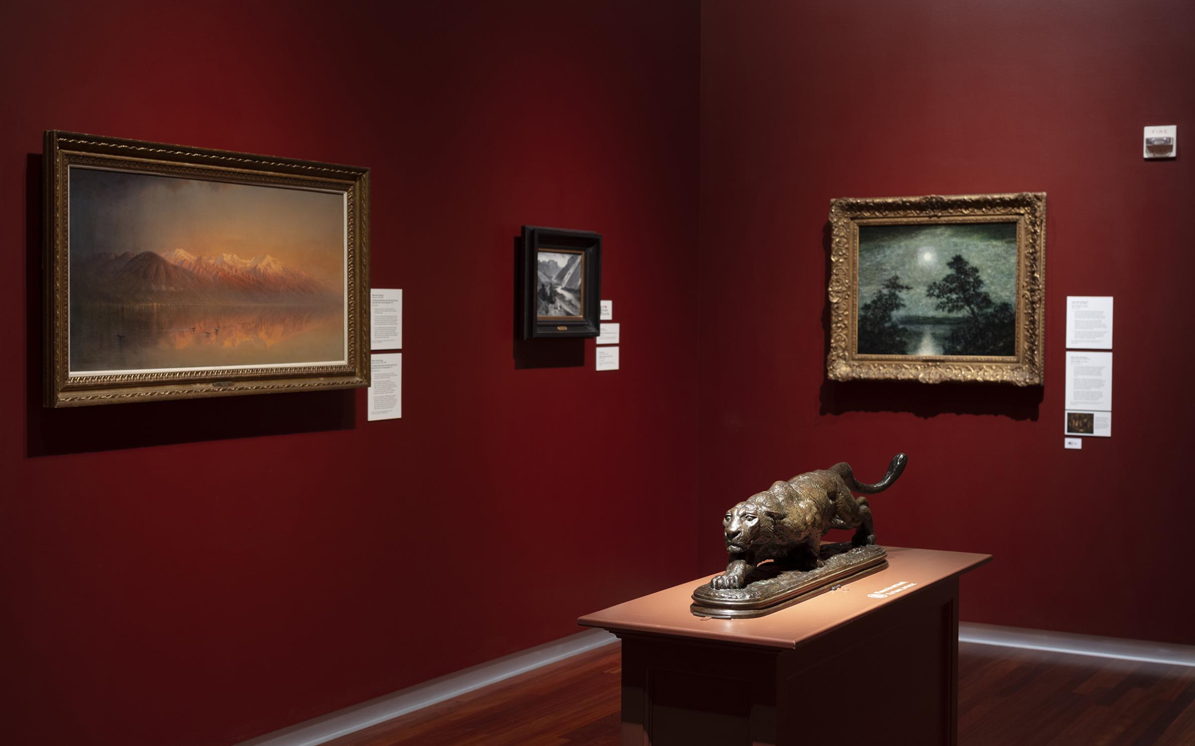 A bronze jaguar statue stands on a mount in the middle of a room with dark red walls with paintings hanging around.