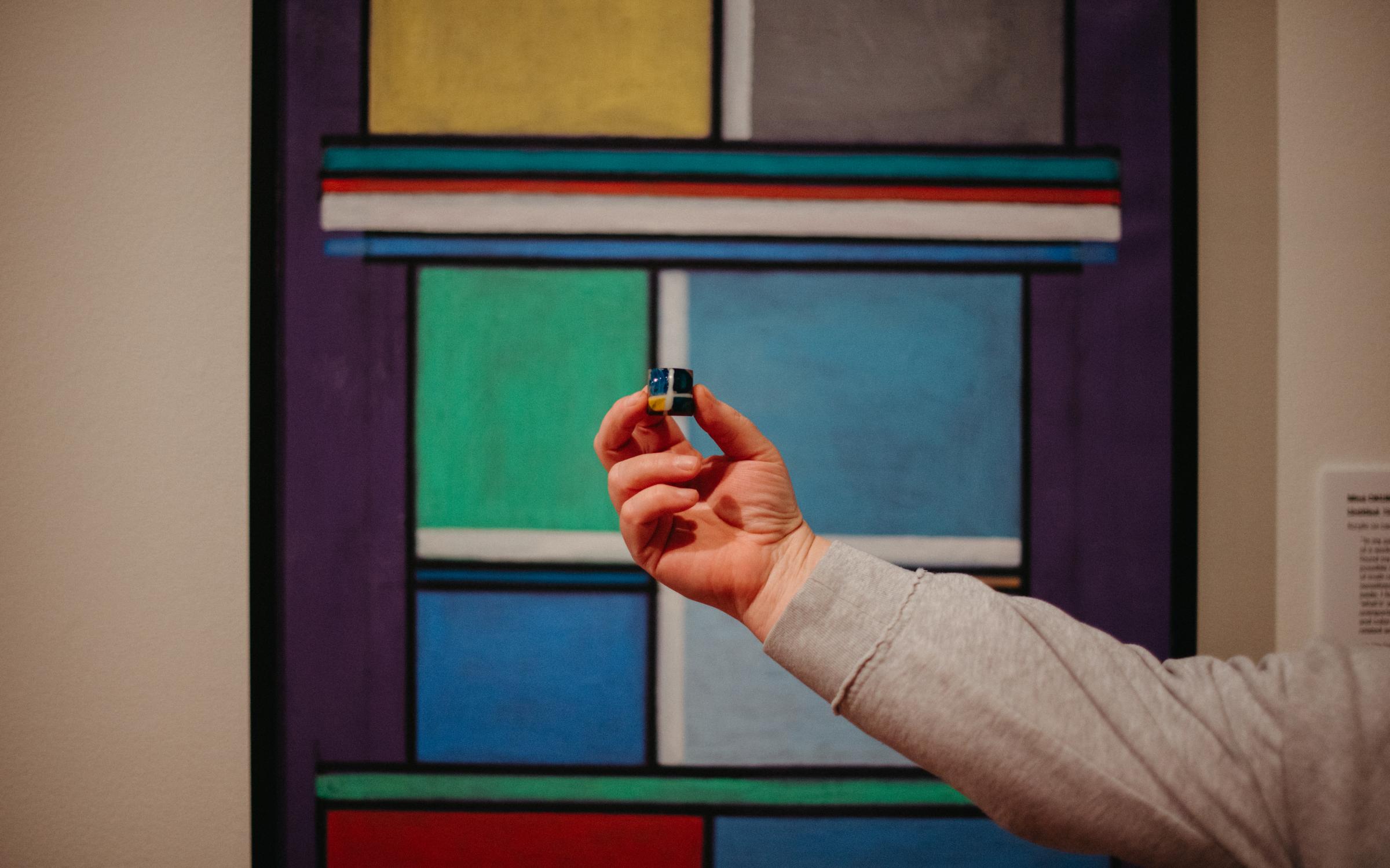 A hand holds a colorful chocolate in front of a color-block painting of the same pattern.