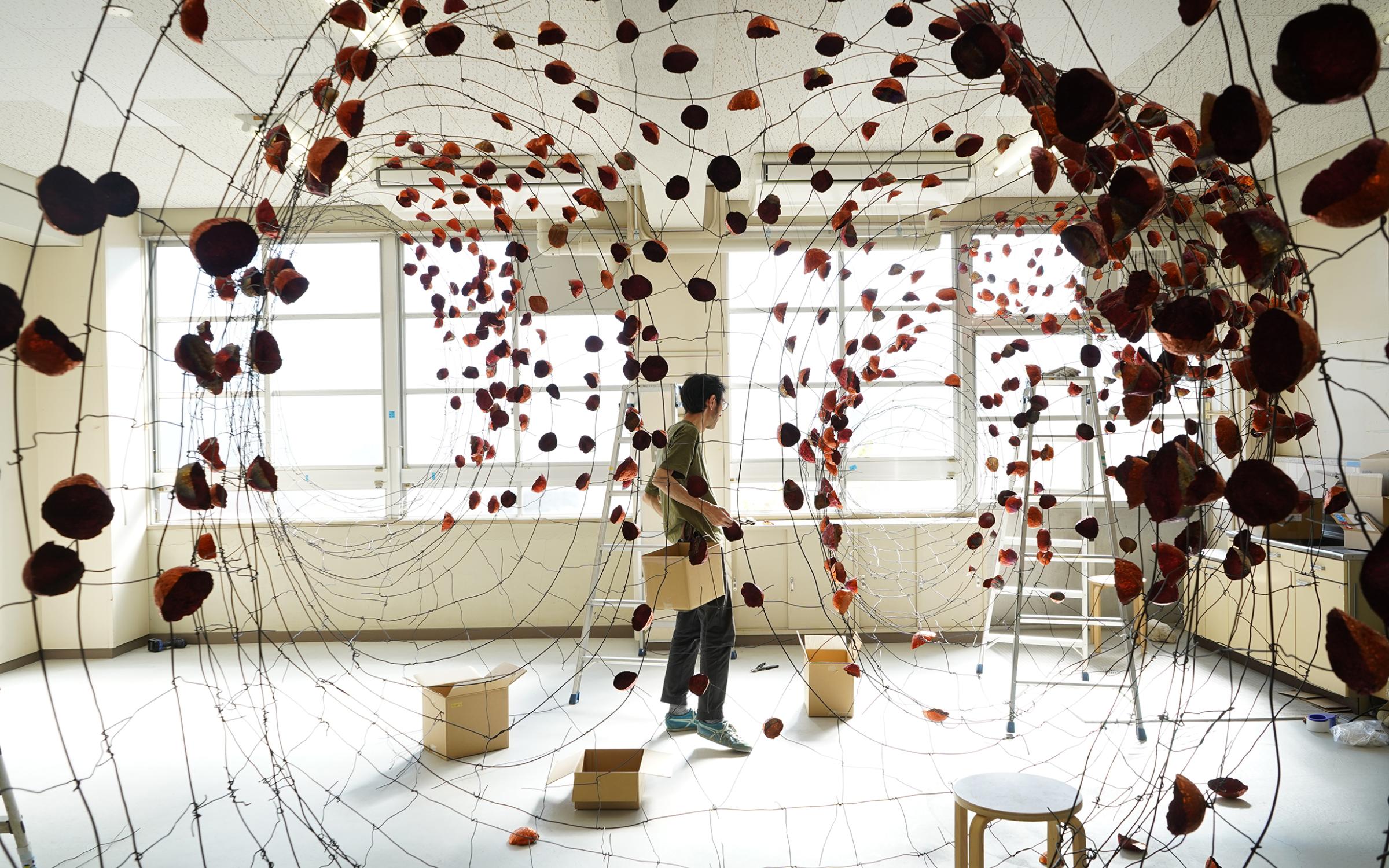 A man stands in the middle of a room with boxes, stools and ladder and small rock shapes copper pieces hanging from wire spread across the room.