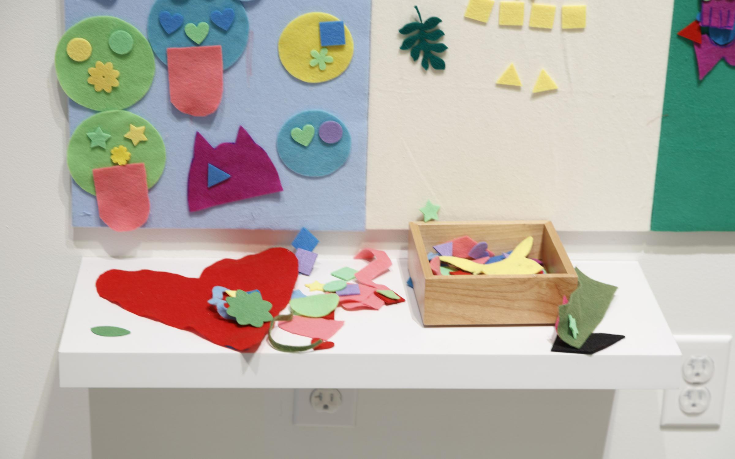 A table covered in felt shapes. Behind it there is a blue and white felt board with colorful felt shapes arranged on it.