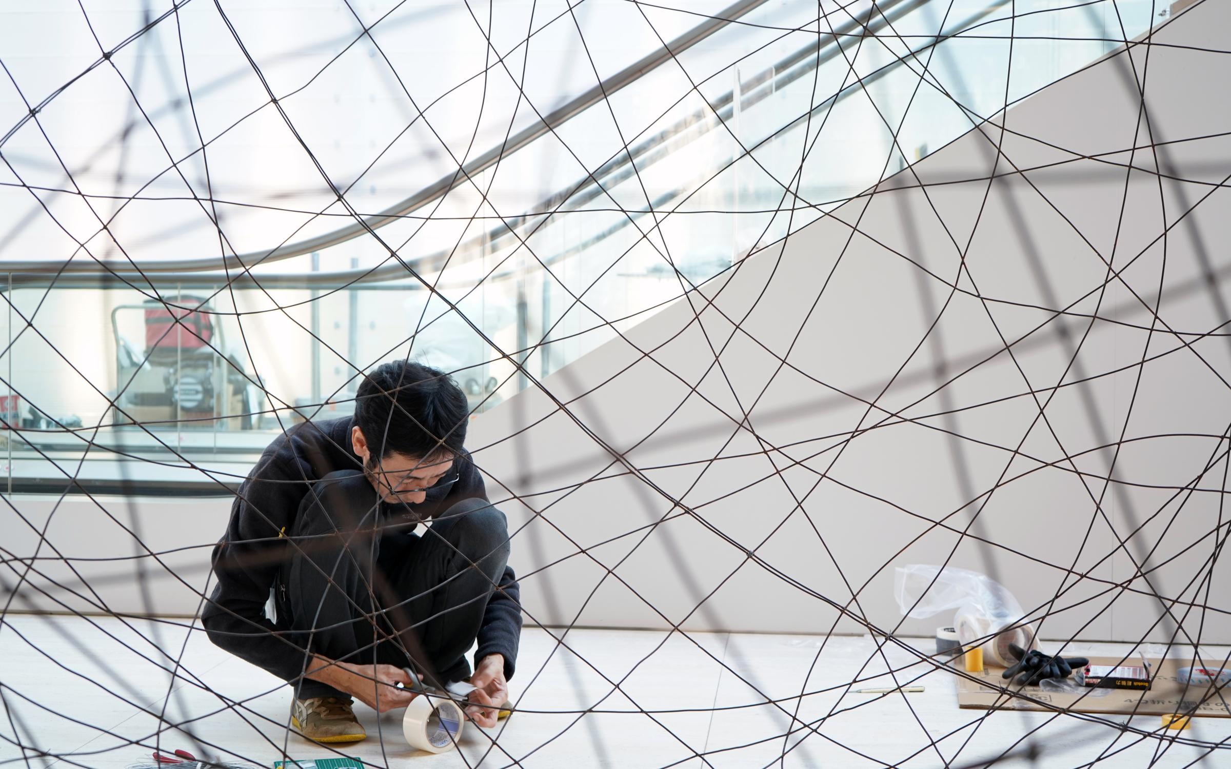 Artist, Minako Yoshida kneels down to work. He is surrounded by wire.