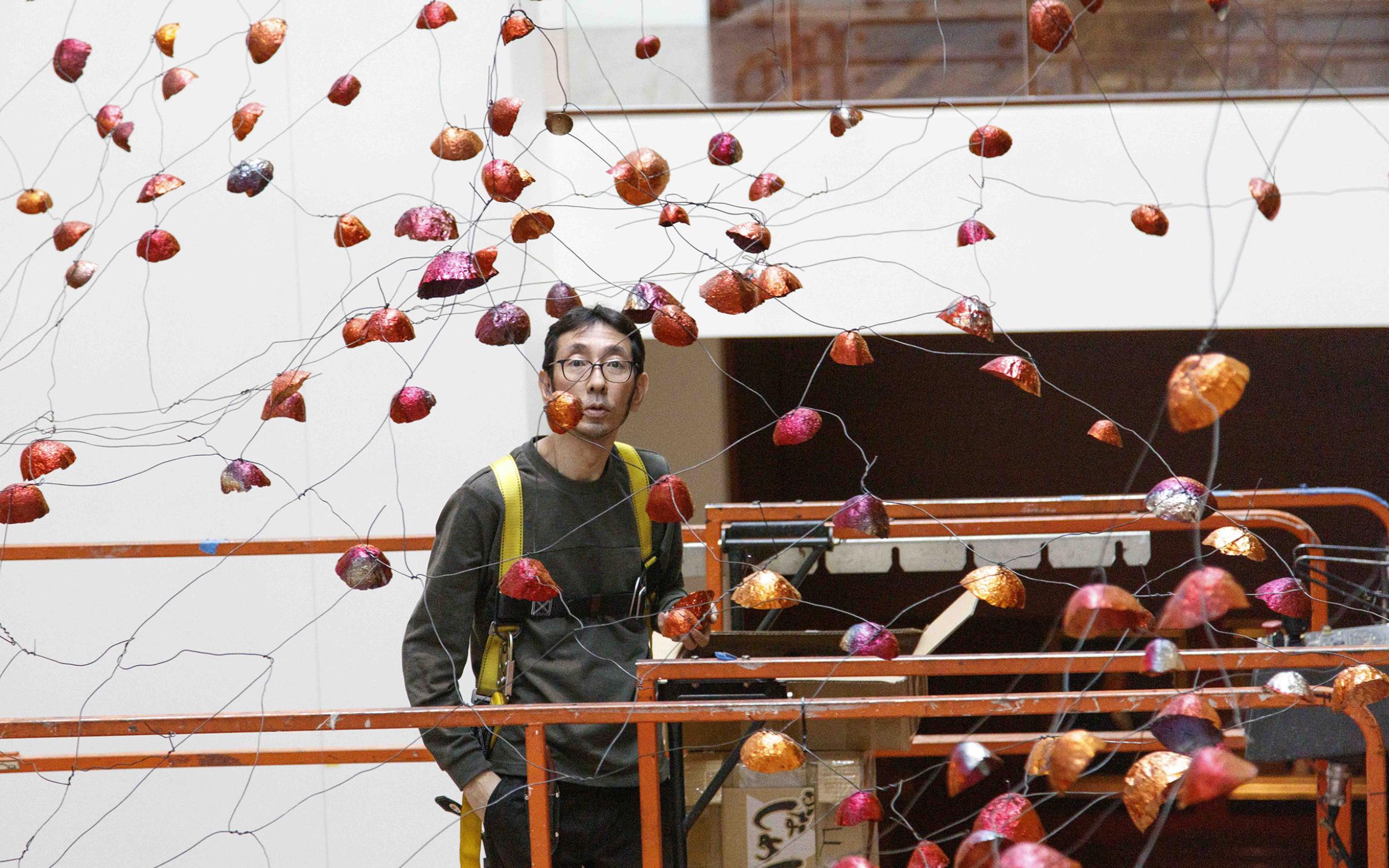 A man stands on a lift in the middle of an art installation of wires and foil, copper cups.