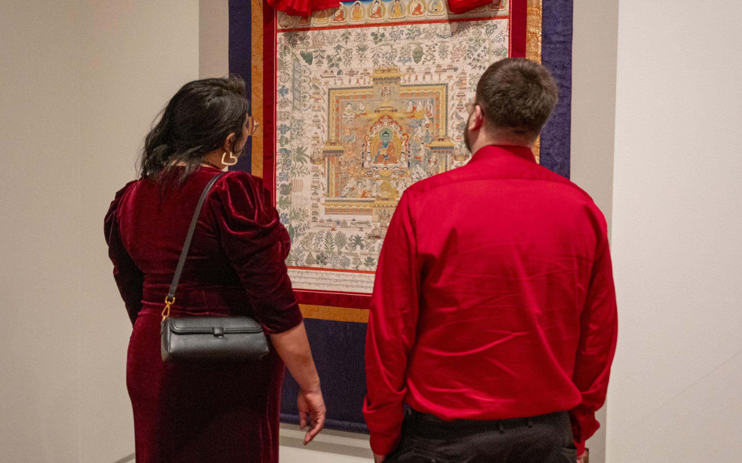 A woman in a long red dress and a man in a long sleeve shirt stand together looking at a Himalayan Art on the wall.