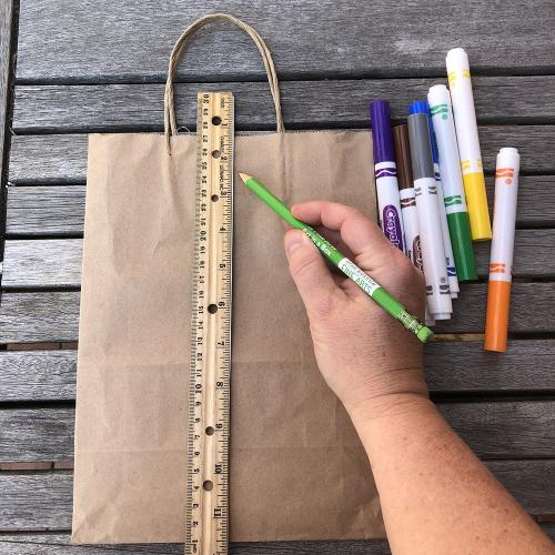 a hand tracing a straight line with a ruler on a paper bag with a green pencil, a pile of colored markers sits next to it