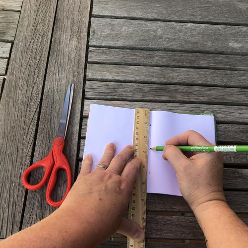 two hands holding a green pencil and a ruler measure down the center of a white sheet of paper a pair of red handled scissors sit on the wooden table top next to the paper. 