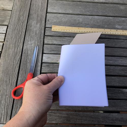 a hand holds a piece of paper folder in half on the short side, a ruler and scissors sit on the table top behind