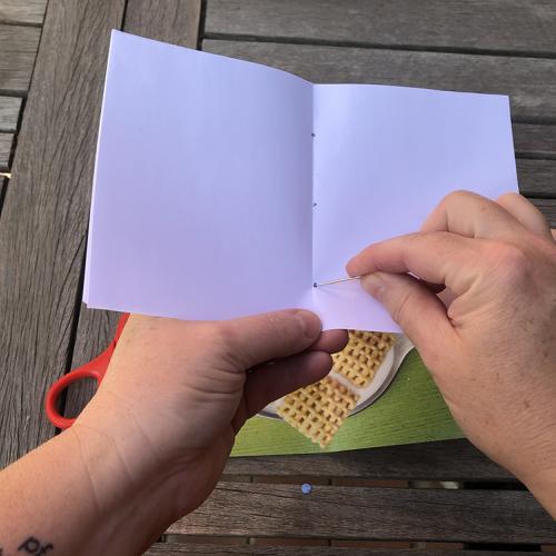 hands holding a sheet of white paper using a needle to poke holes on a center fold