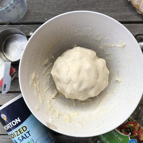 a white bowl with a ball of fresh salt dough in it is surrounded by salt dough supplies, salt, oil, measuring cup and measuring spoons 