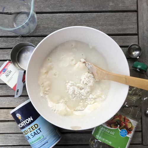 a white bowl with a wood spoon and flour water in it is surrounded by salt dough supplies, salt, oil, measuring cup and measuring spoons 