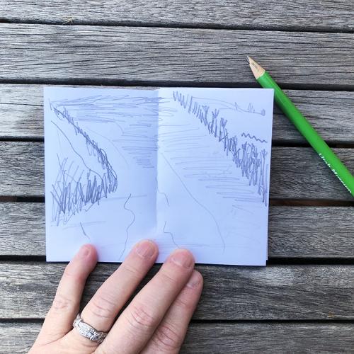a white sketchbook with a pencil drawing of a stream with a green pencil on the table 