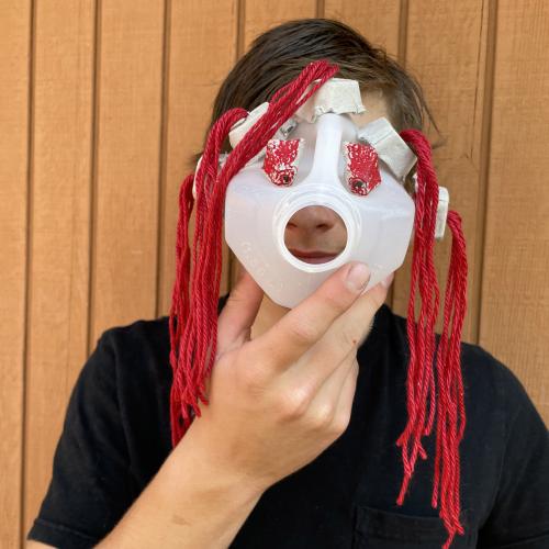 a child in a black t-shirt holding up a mask made from a milk carton and red yarn