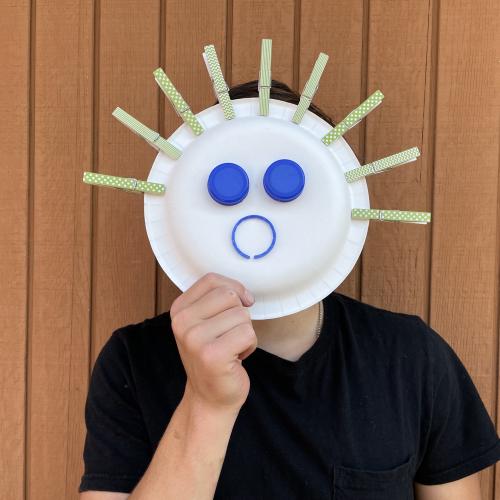 a child in a blak t-shirt holding up a mask that made from a paper plate that looks like it has a surprise face on it