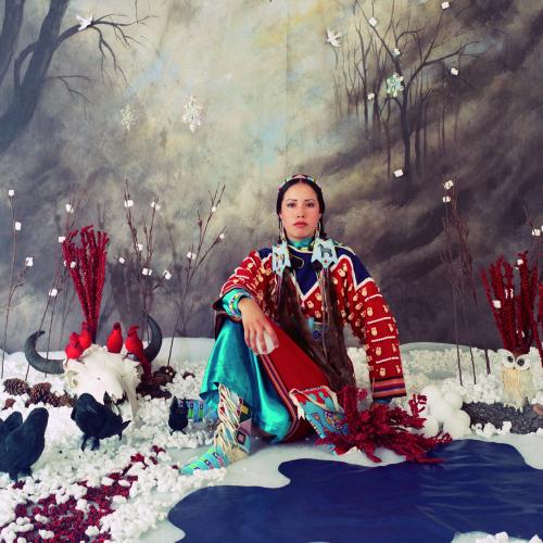 an indigenous women wearing traditional dress of a red coat adorned with white bones and beads and a turquoise blue skirt, sit in a winter scene built in a studio, the is fake snow and a painted backdrop and bison skull with four fake red cardinals on it sits next to her, the is also a group os fake crows and a a fake owl. 