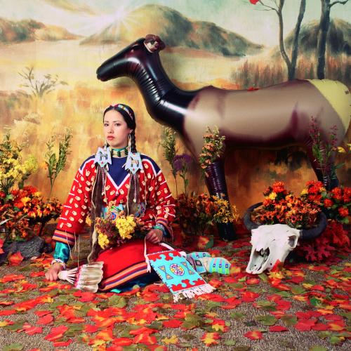 an indigenous woman sits in a fall scene composed in a studio. She is wearing red traditional dress adorned with white bones and beads. there is a painted backdrop of a yellow grassin hill and an inflatable elk next to a bison skull. the ground is covered in fake leaves.  