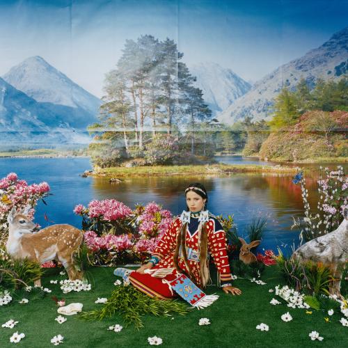 an indigenous women wearing traditional dress of a red coat adorned with white bones and beads and a turquoise blue skirt, sits in a springtime scene composed in a studio. There is a painted backsrop of a placid mountain lake in the background. The Foreground consists of the woman sitting on fake grass strewn with white flowers, there is a stand of pink peonies behind her, cardboard cutouts of a doe, rabbits and a coyote fill out the scene.