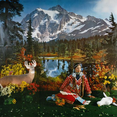an indigenous women wearing traditional dress of a red coat adorned with white bones and beads sits in a summertime scene composed in a studio. There is a painted backdrop of a placid mountain lake surrounded by pine forrest in the background. The Foreground consists of the woman sitting on fake grass with yellow and orange fake flowers surrounding her, there is a cardboard cutout of a buck on the side of the scene to the left and a cutout of a rabbit and a white plastic cow skull on the ground to the righ 