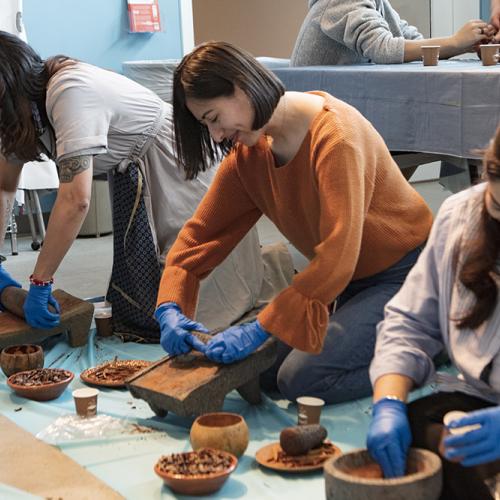 Three women with dark hair kneel on blue plastic squares on the floor. They wear blue gloves and are grinding cacao seeds.