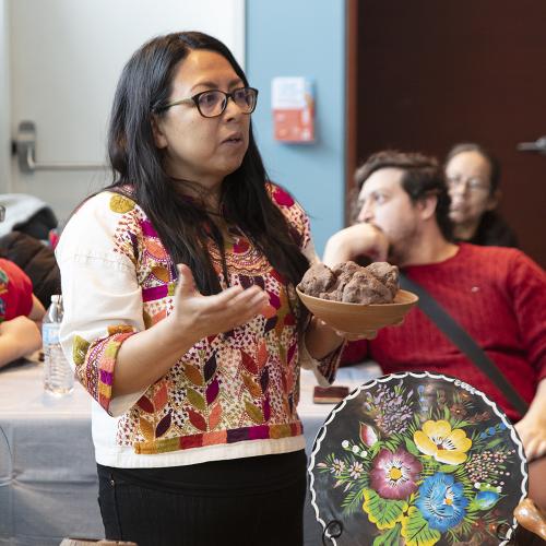 A woman with long, dark hair holds a bowl in her left hand while talking in the middle of a room full of adults. She is wearing a colorful shirt. There's a colorful, floral piece of art on the table next to her.