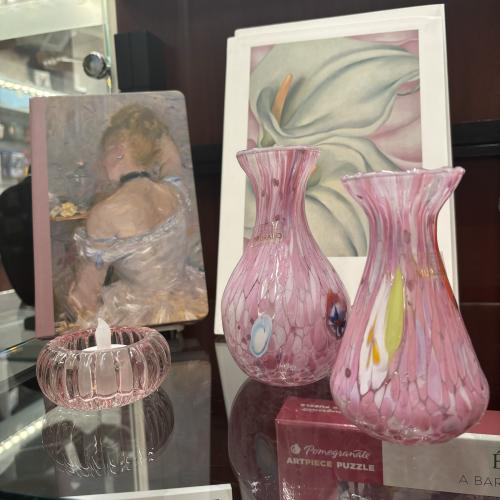 pink Murano glass vases on a glass table in front of a painting of white lily and a pink book with a white ballerina with blonde hair.