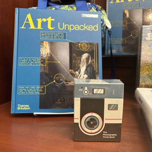 a book with a blue cover titled "Art Unpacked" sits behind a deck of cards in a plastic-wrapped box that looks like a camera.