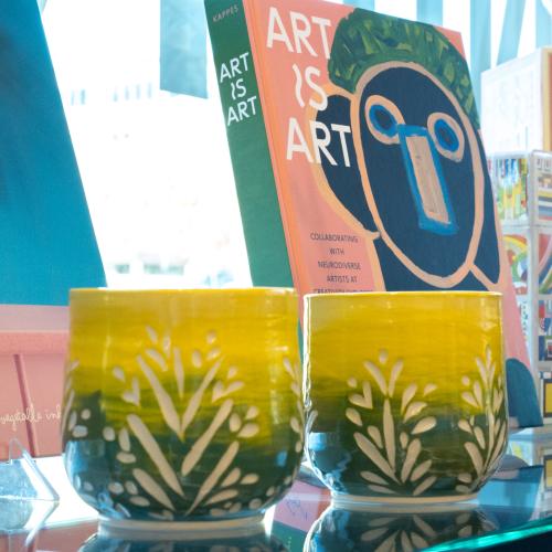 Two large, green and yellow, ceramic mugs without handles sit in front of a book titled, "Art is art".