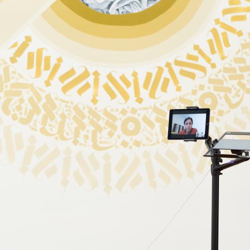 A screen with someone on a video-call sits in front of a mural in-progress.