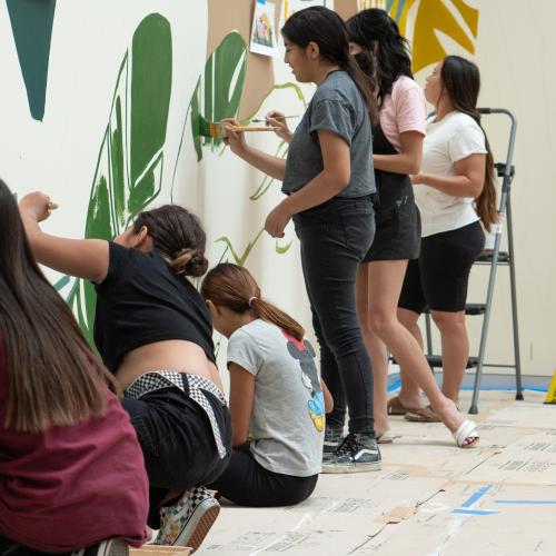 Young people sit and stand on the floor painting the base of a large mural.