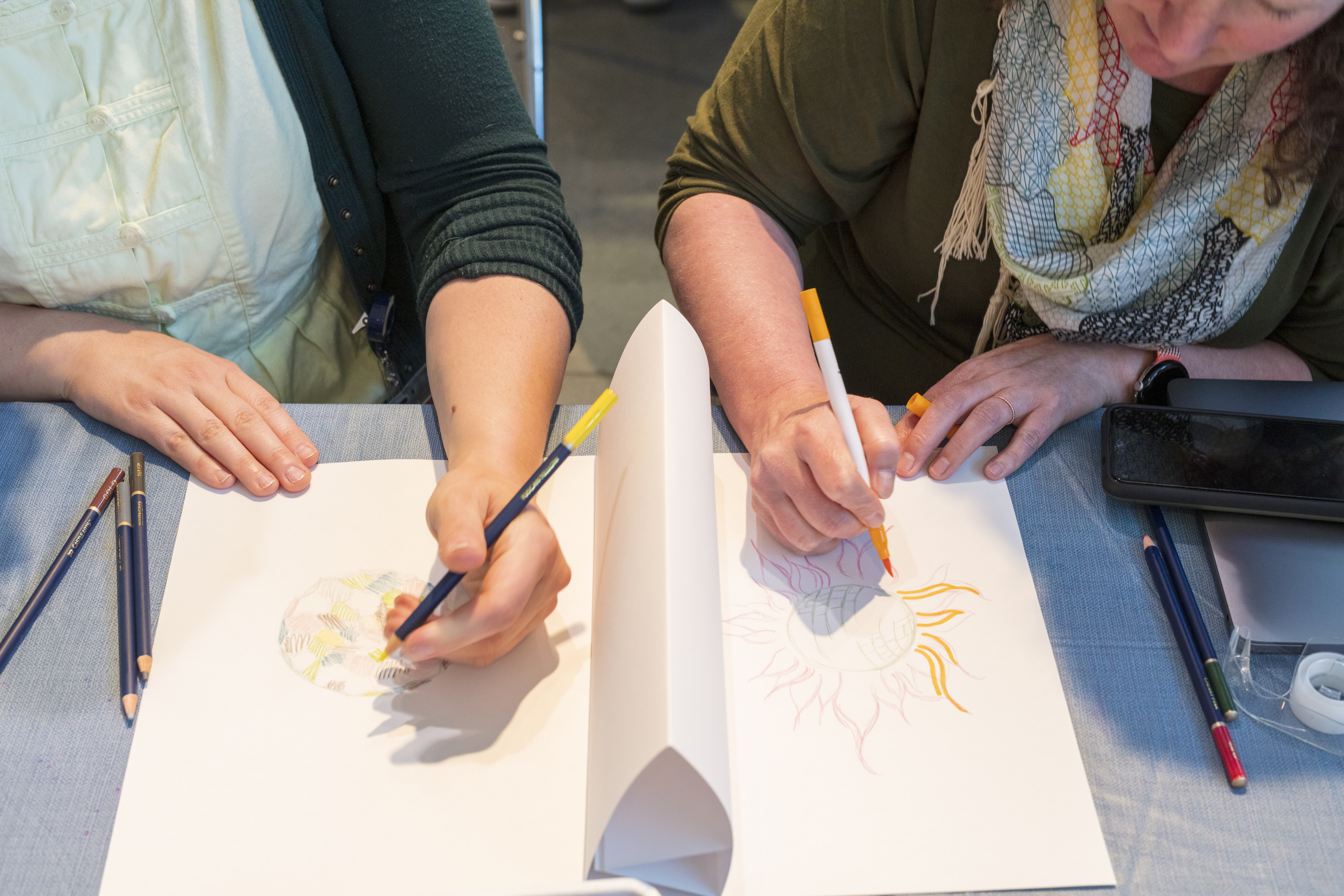 A pair of people draw on halfs of paper with a folded triangle sheet between them.