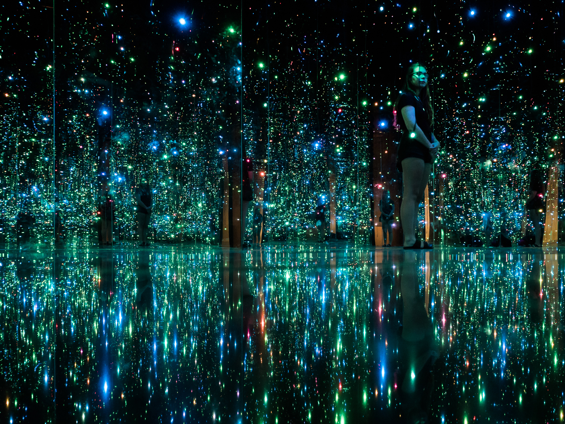 A dark room filled with colorful lights, like fireflies. The room has mirrors on the floor and walls and seems to go on for eternity.