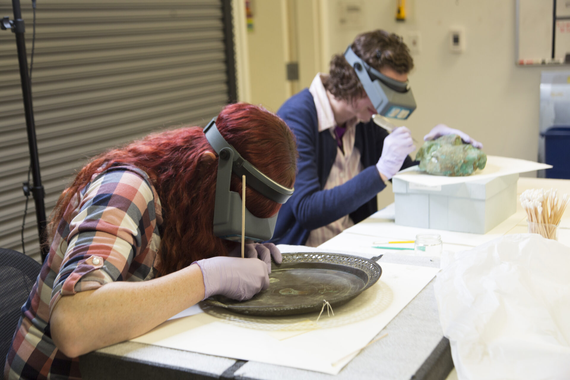 Two people wearing magnifying headbands and gloves carefully clean and examine artifacts, including a large metal tray, at a table in a conservation setting.