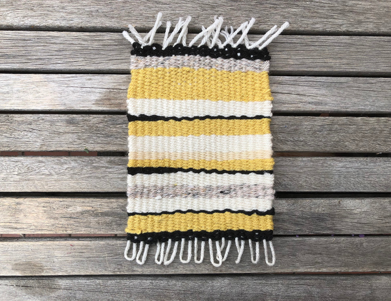 A small woven rug on a wooden background. The rug has black, yellow, beige, and white stripes of different thicknesses.