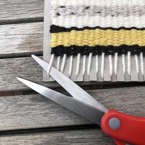 Scissors cutting the bottom of vertical strips of white yarn hooked to a piece of cardboard
