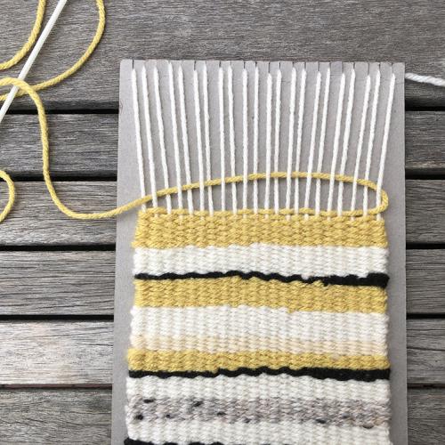 Yellow yarn being woven back the opposite direction above and underneath the vertical yarn on the cardboard