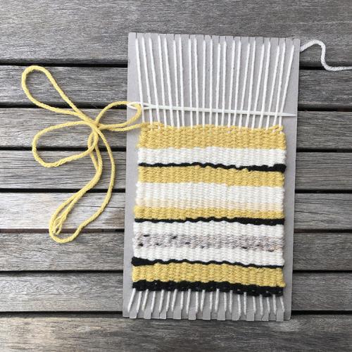 Yellow yarn threaded through a long plastic needle that is being woven in and out of the vertical strips of yarn already attached to the cardboard.