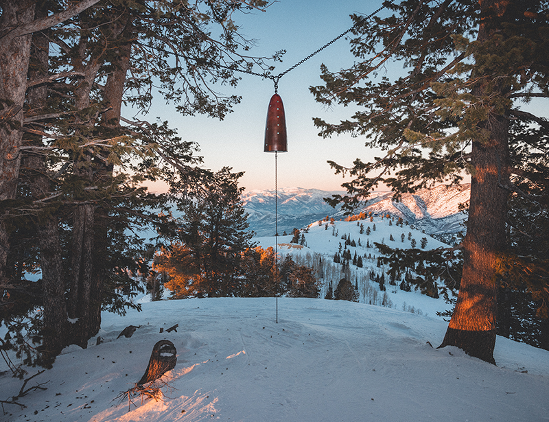 A bell strung between two trees on a snowy hill.