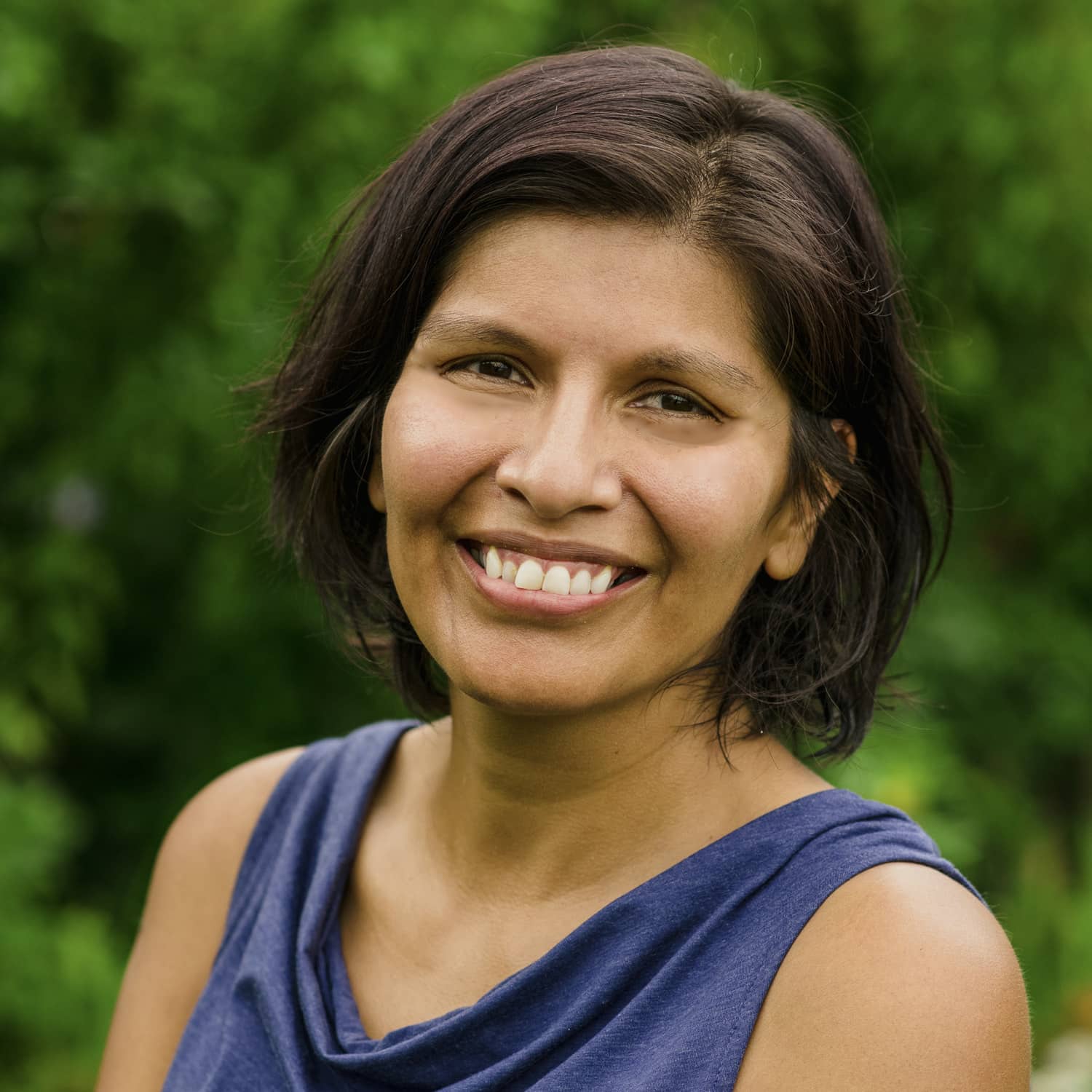 A woman with short dark hair and a sleeveless blue top smiles warmly at the camera, standing outdoors with a blurred background of lush green foliage.