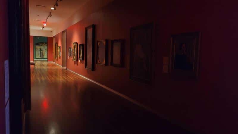 A dimly lit art gallery hallway with framed paintings on red walls, polished wooden floors, and a softly illuminated doorway at the far end.