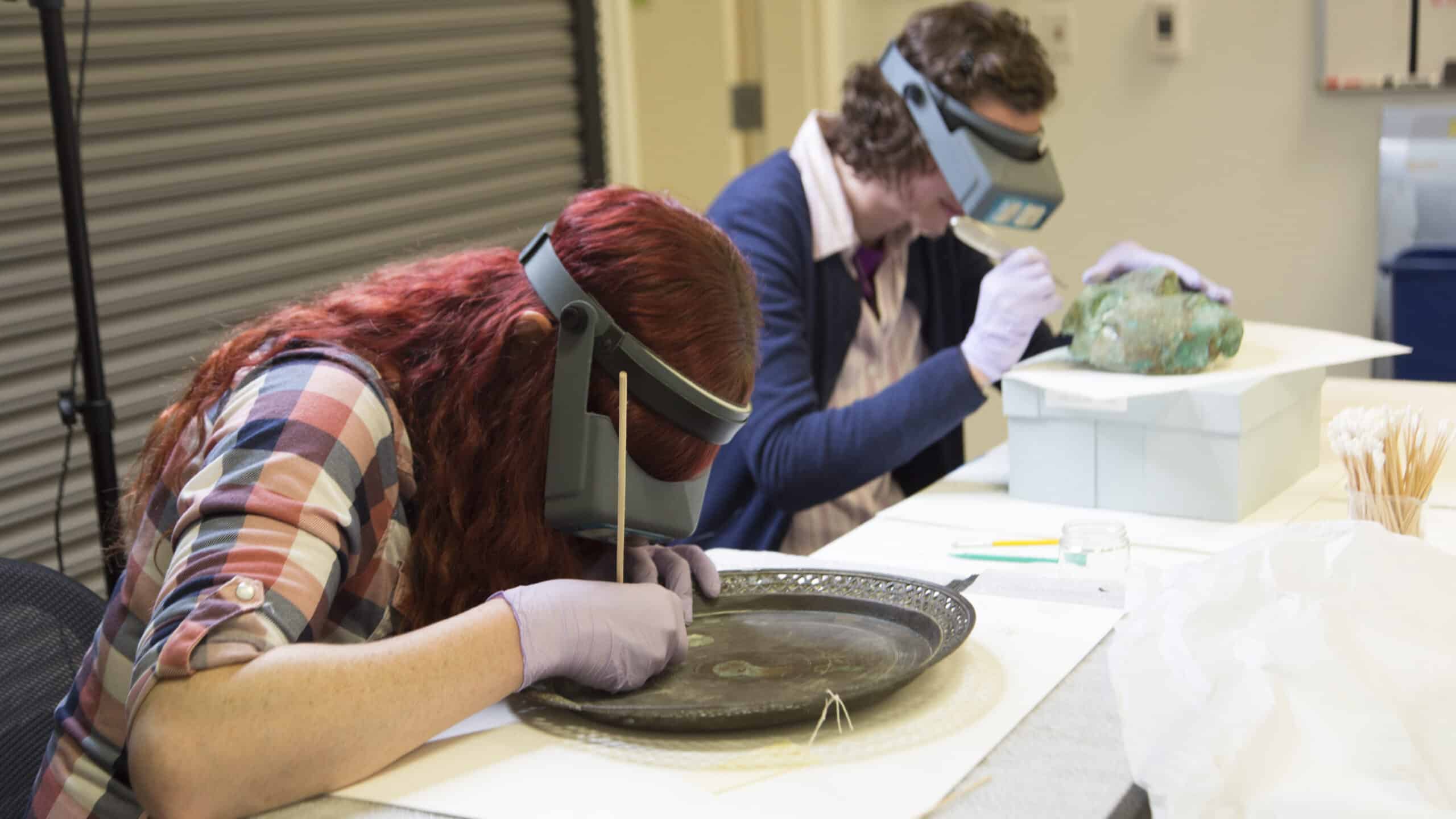 Two people wearing magnifying headbands and gloves carefully clean and examine artifacts, including a large metal tray, at a table in a conservation setting.