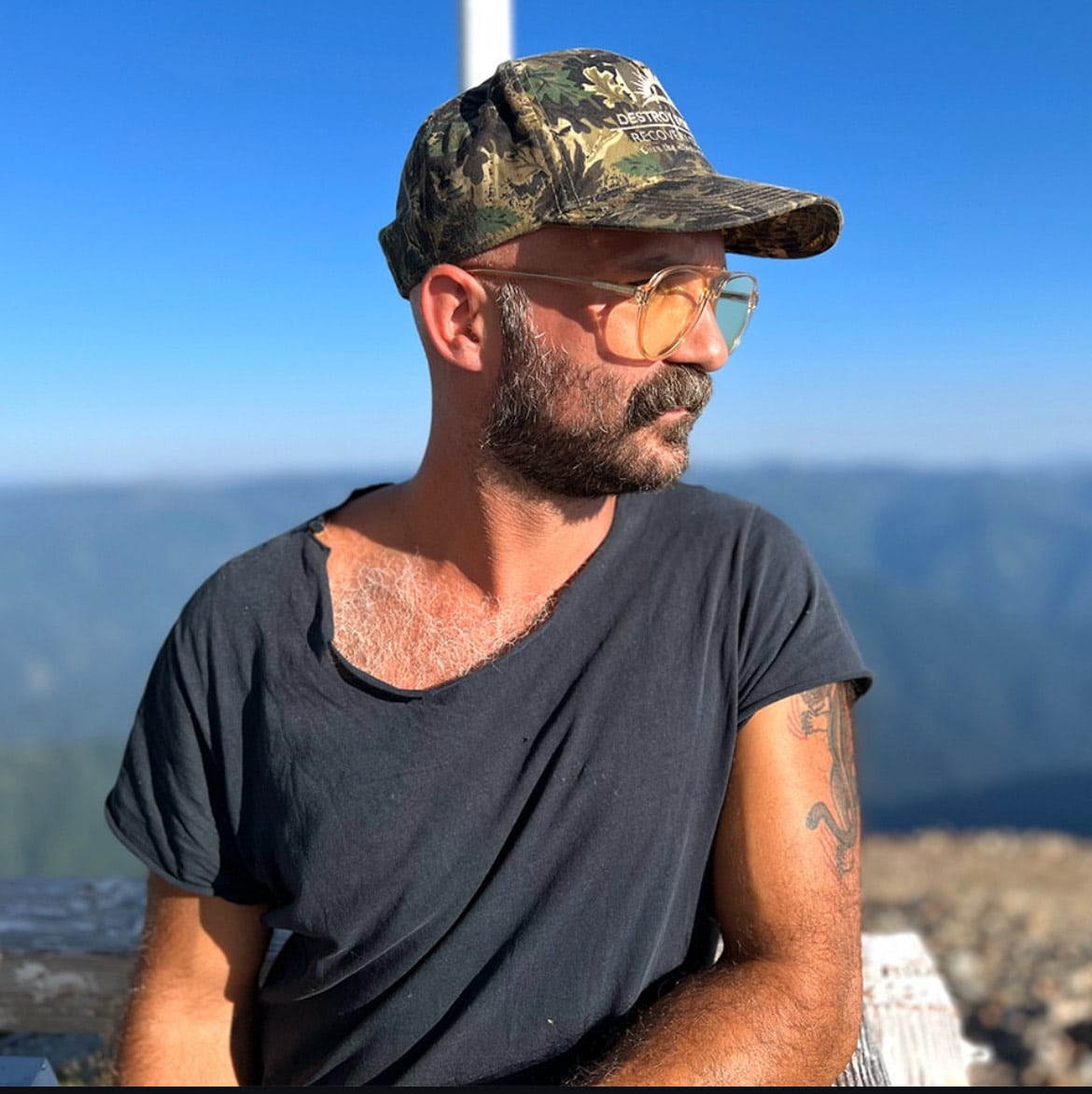 A man, Jacob Riddle, with a mustache and beard wears a camouflage cap, large glasses, and a ripped black T-shirt, sitting outdoors with mountains and a blue sky in the background.