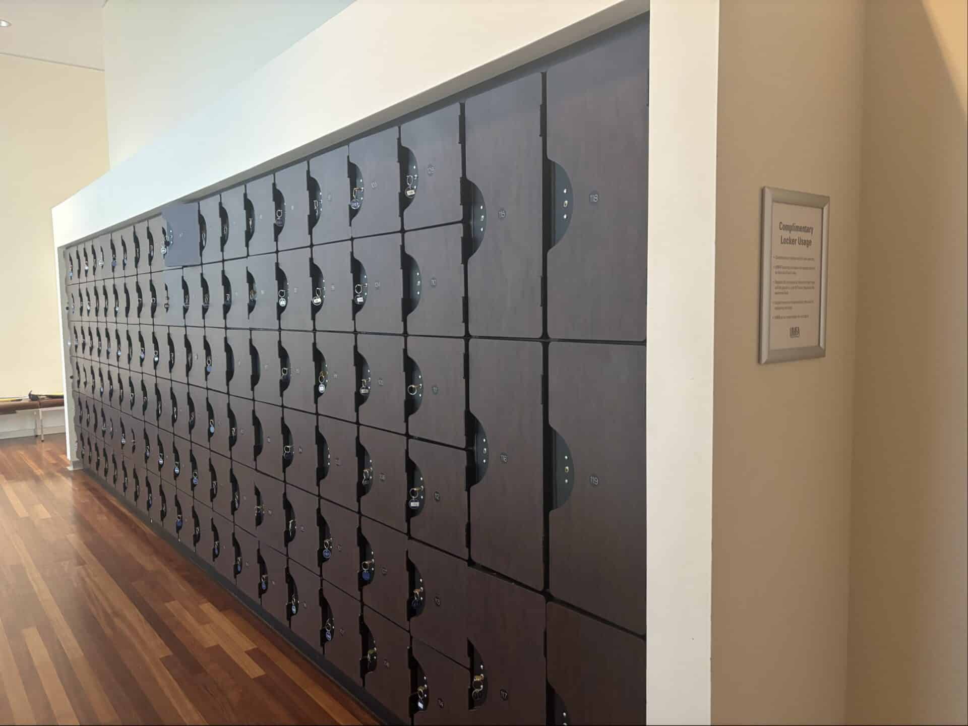 A long row of dark wooden lockers with silver locks is positioned against a white wall in a room with polished wooden floors. A sign is mounted on the wall beside the lockers.
